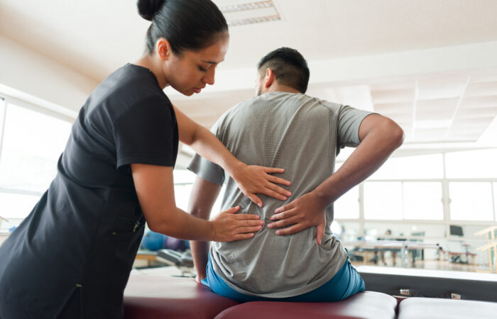 A female massage therapist standing behind a male patient and massaging his back with both hands.